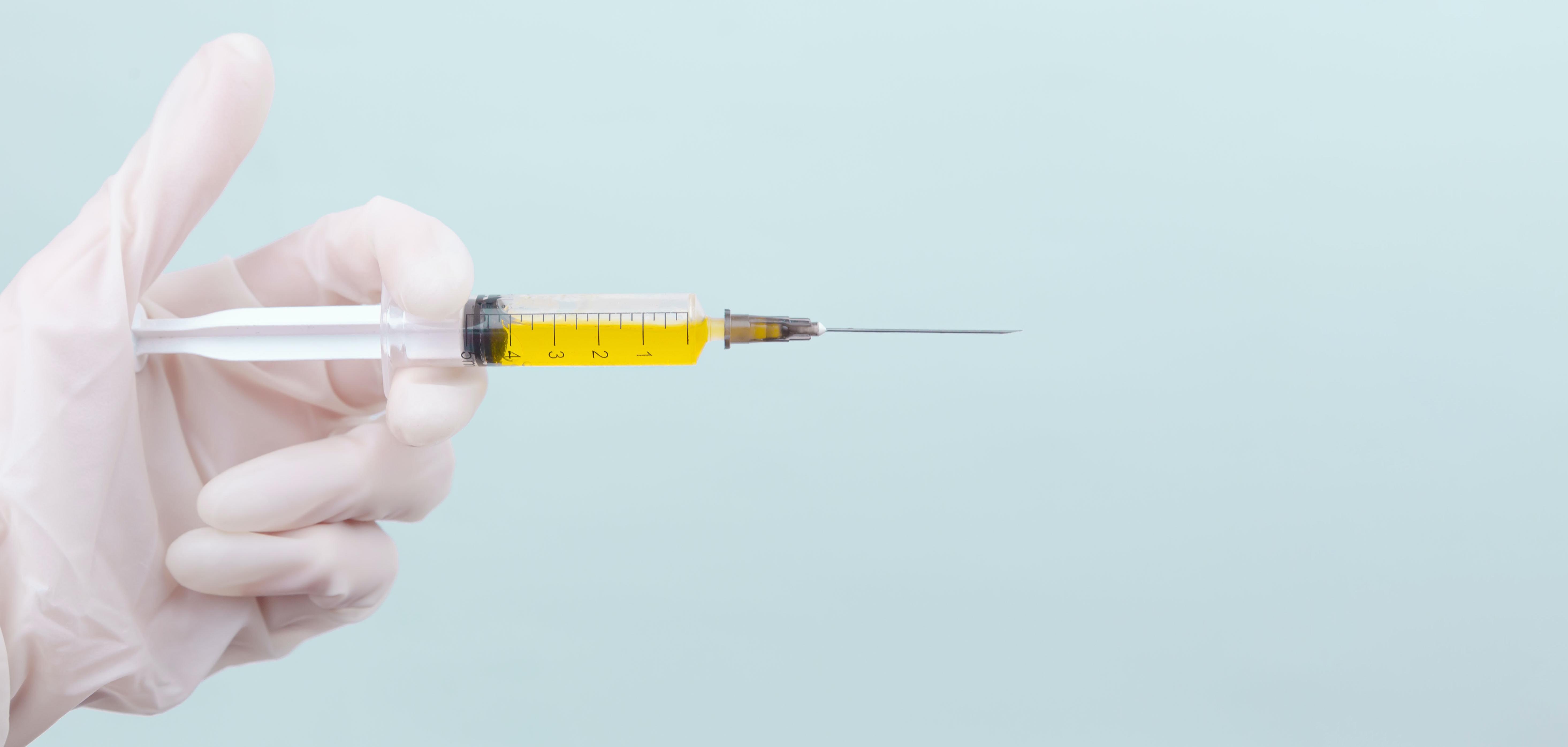 The doctor's hand in a white medical glove holds a syringe with a yellow medicine on a blue background.